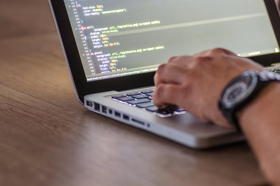 This image shows a person working on a laptop placed on a wooden desk, with visible code on the screen. The individual is wearing a watch on their left wrist and is using their right hand to type or n